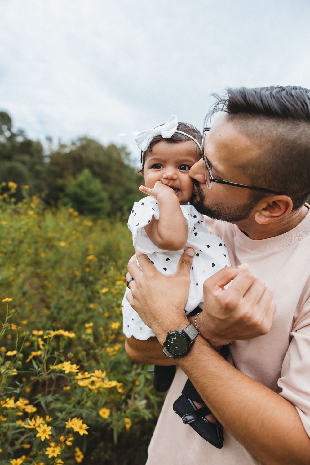 Father kissing baby girl in white polka dot dress with bow headband outdoors among yellow wildflowers.
