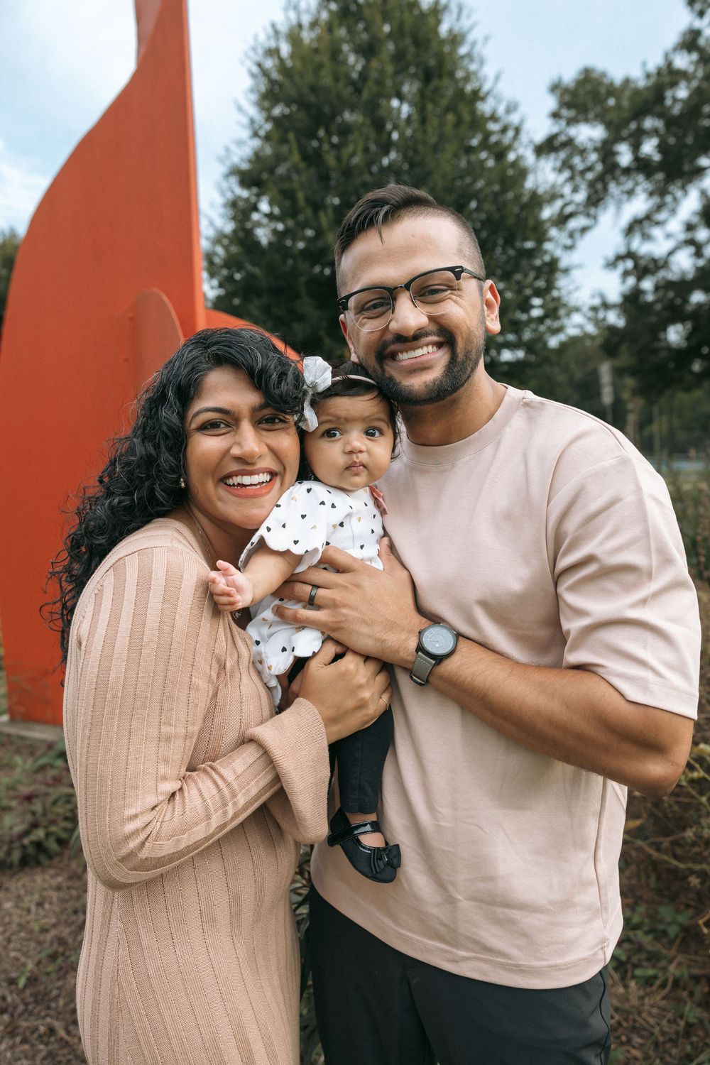 Smiling family with parents holding their baby girl outdoors near a red sculpture and green trees.