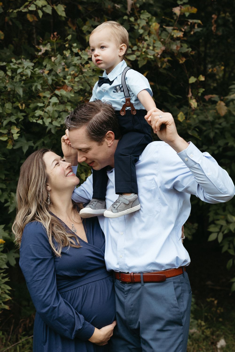 Pregnant woman in blue dress with husband and toddler son on shoulders posing outdoors among green foliage.