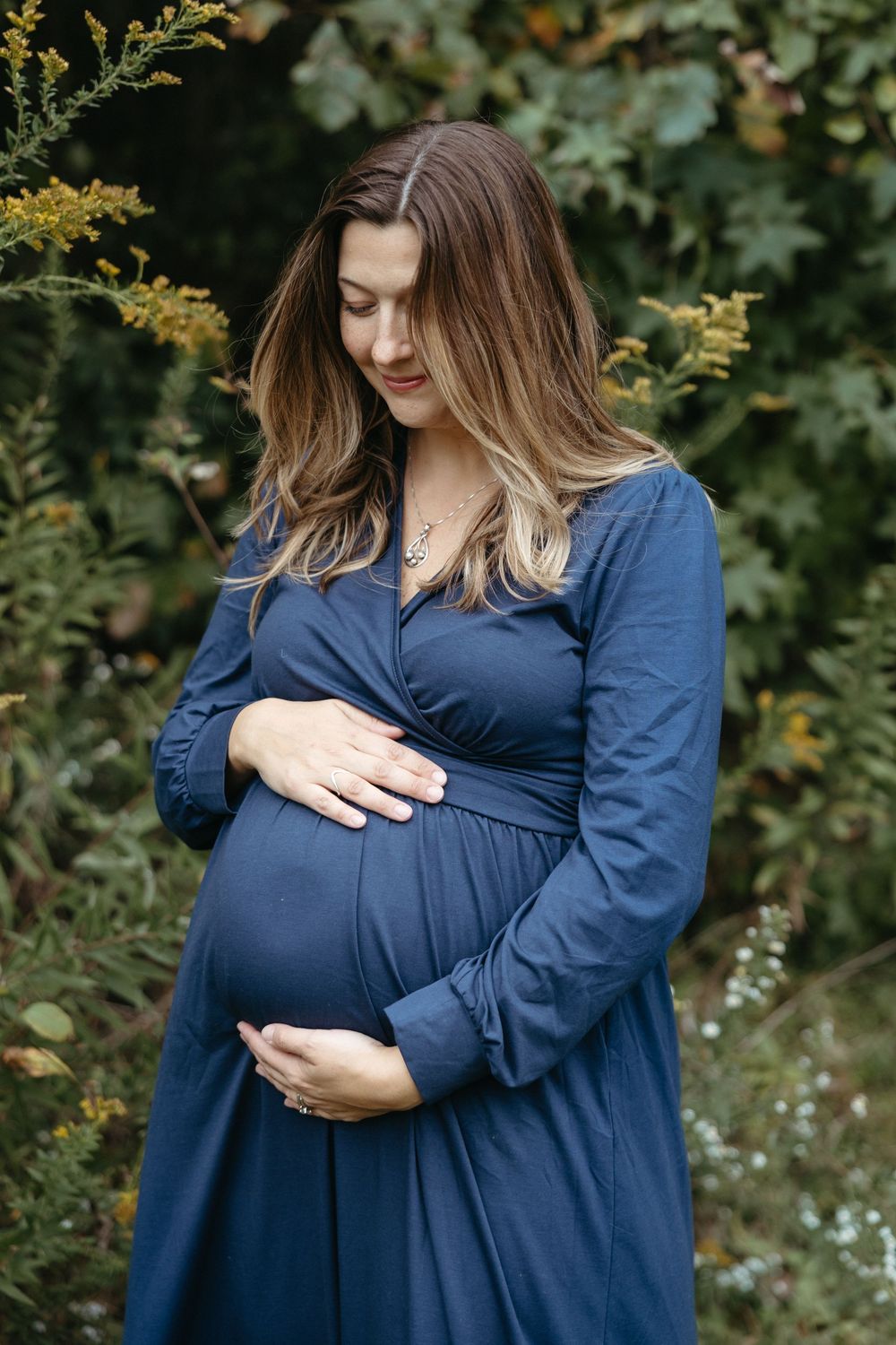 Pregnant woman in navy blue dress cradling baby bump outdoors among green and yellow foliage.