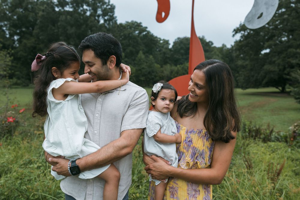 Family of four posing outdoors in a green field near a red sculpture, parents holding two young daughters.