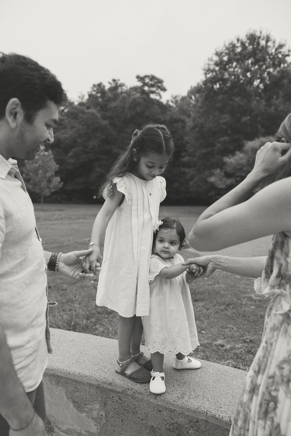Black and white family photo of two young girls in white dresses holding hands with parents in a park.