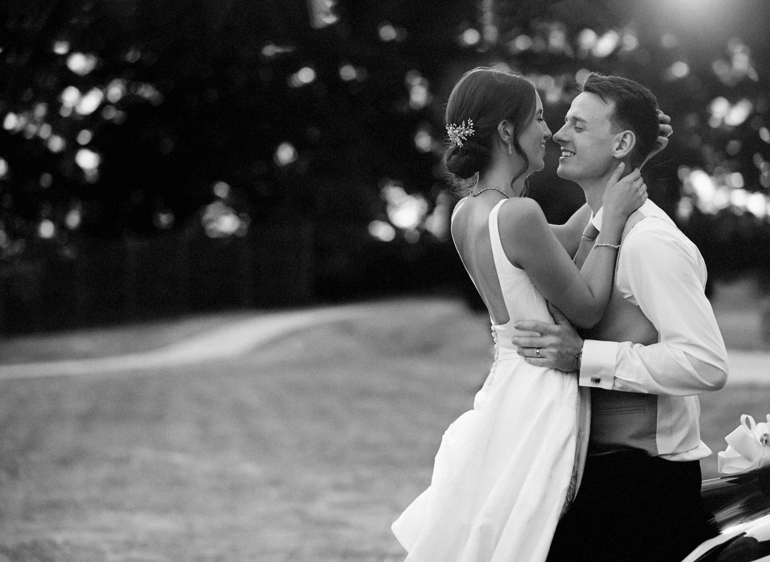 Black and white wedding photo of a couple embracing and smiling outdoors, bride in white dress, groom in white shirt.
