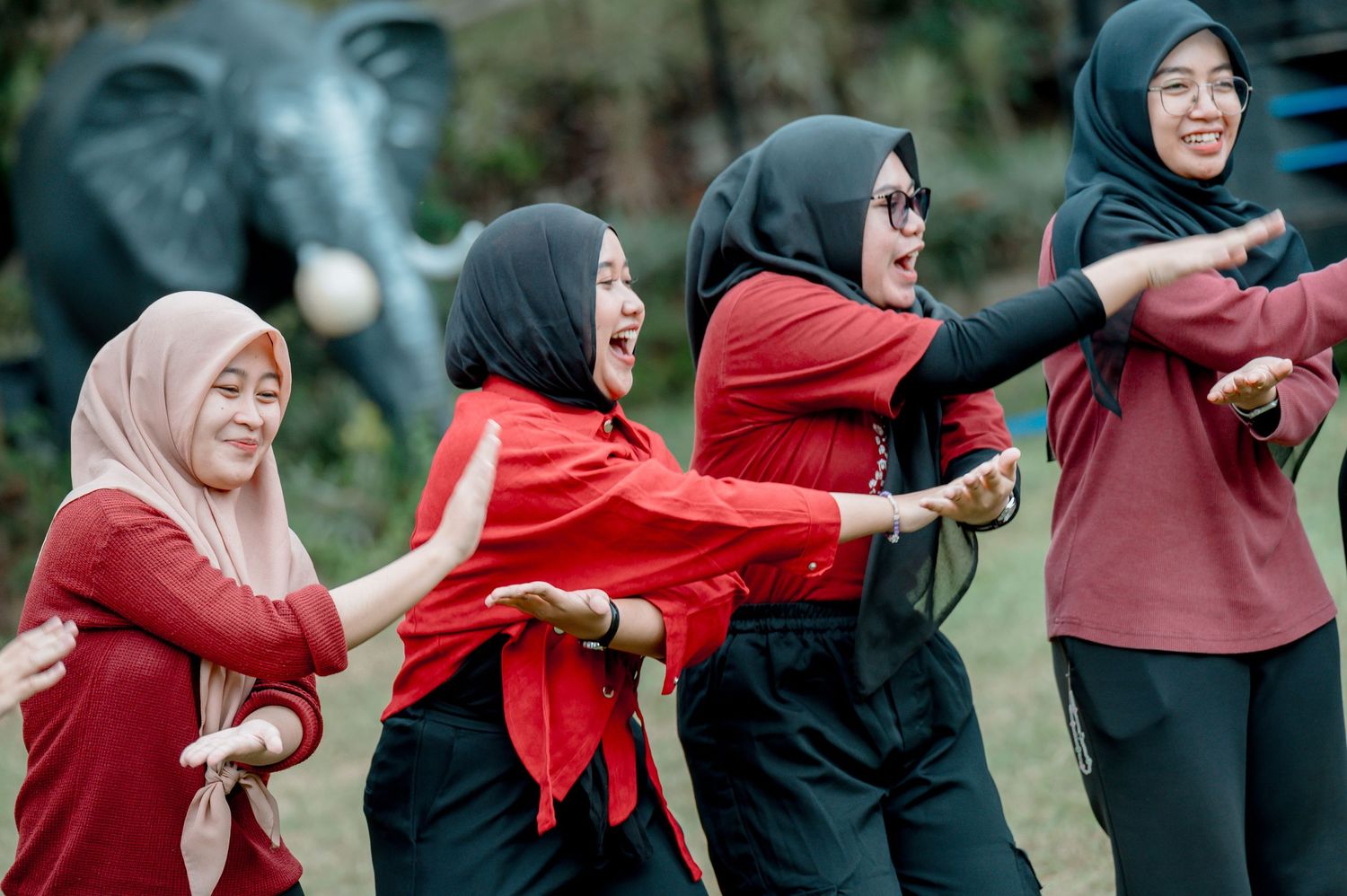Group of women in hijabs wearing red outfits laughing and dancing together outdoors near an elephant statue.
