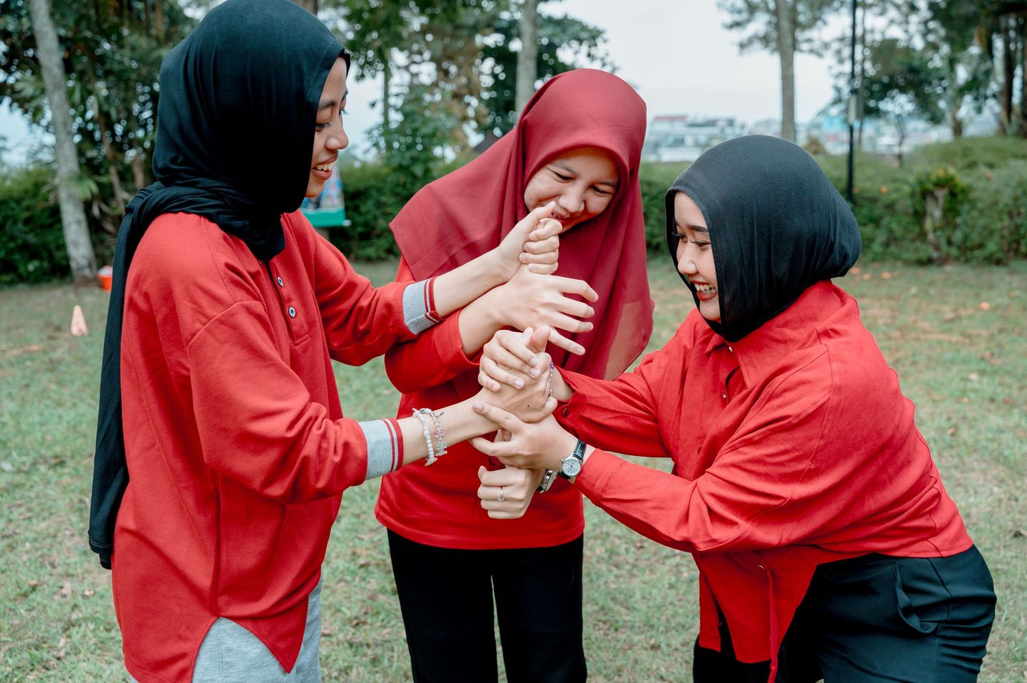 Three women in red shirts and hijabs join hands together outdoors in a team-building exercise.