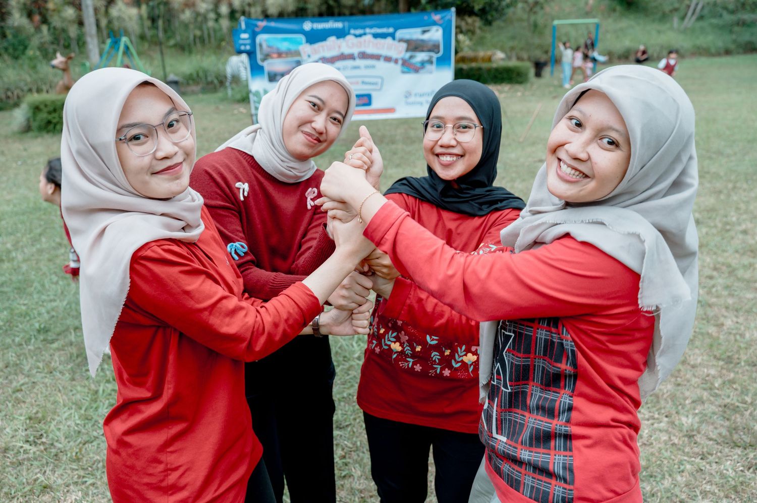 Four young women in hijabs and red tops joyfully join hands together outdoors at a team-building event.