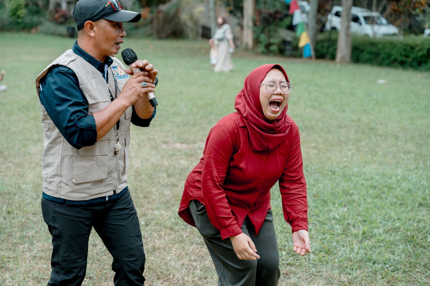 A man holds a microphone while a woman in a red hijab laughs joyfully outdoors in a green park.