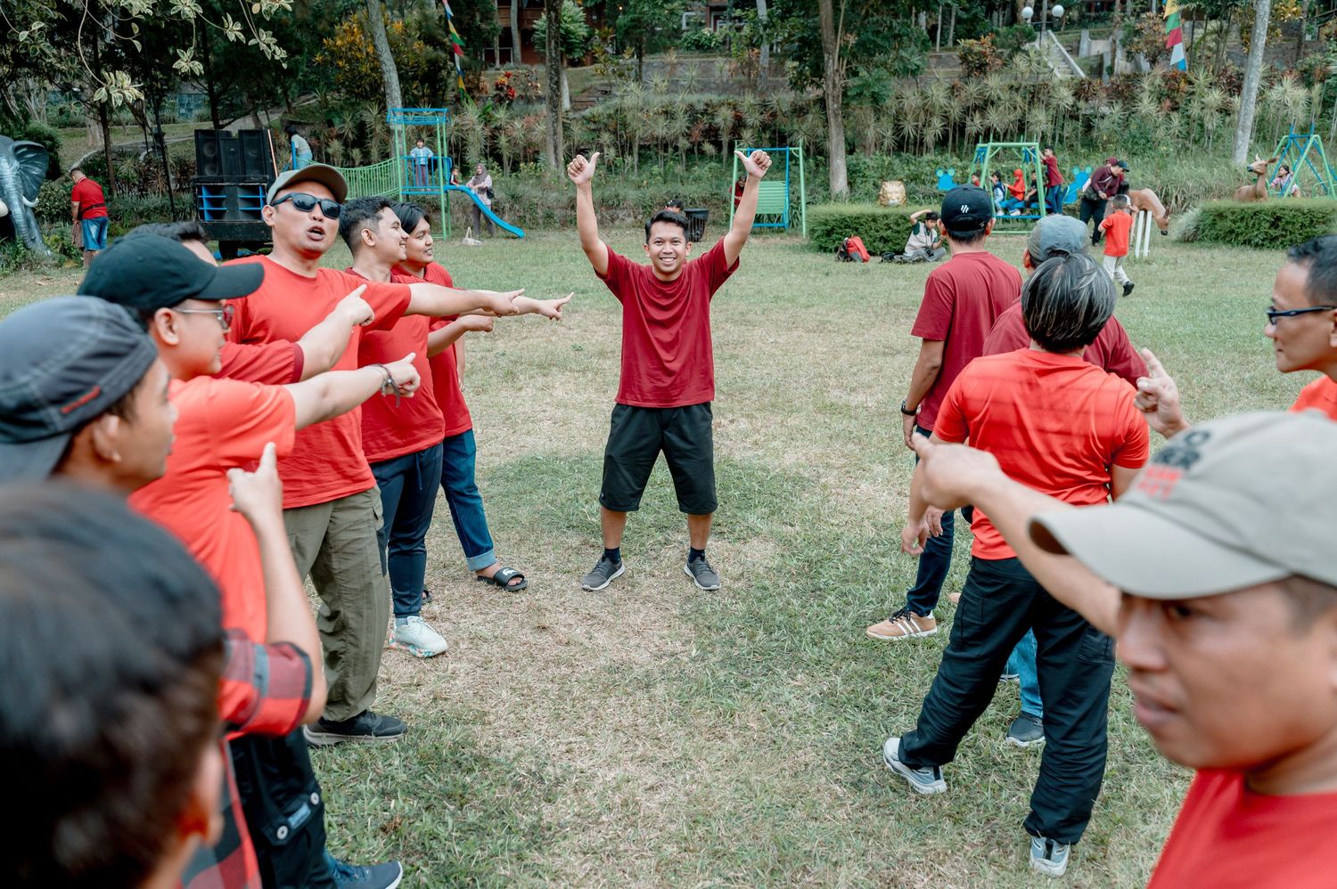 Group of people in red shirts playing outdoor team-building game in a park, led by enthusiastic instructor.