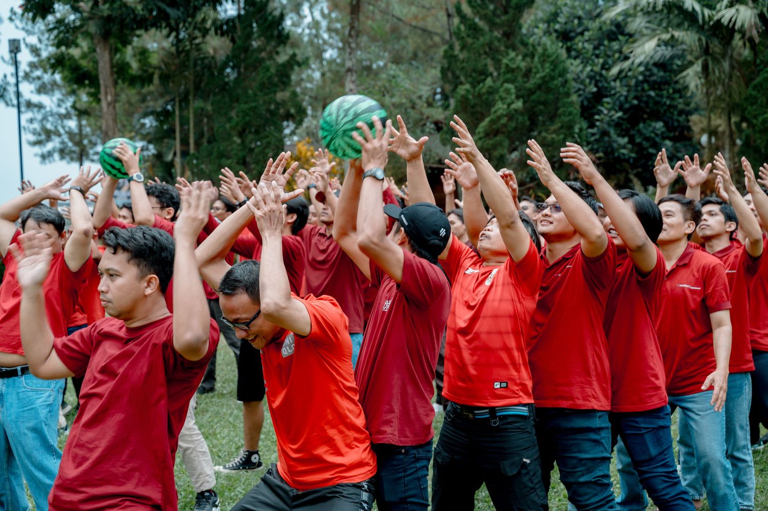 Large group of men in red shirts reaching up to toss a watermelon outdoors during a team-building activity.