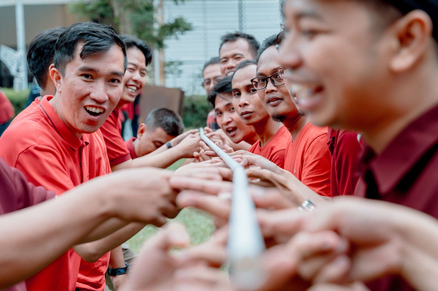 Group of men in red shirts smiling and passing a silver trophy together outdoors.