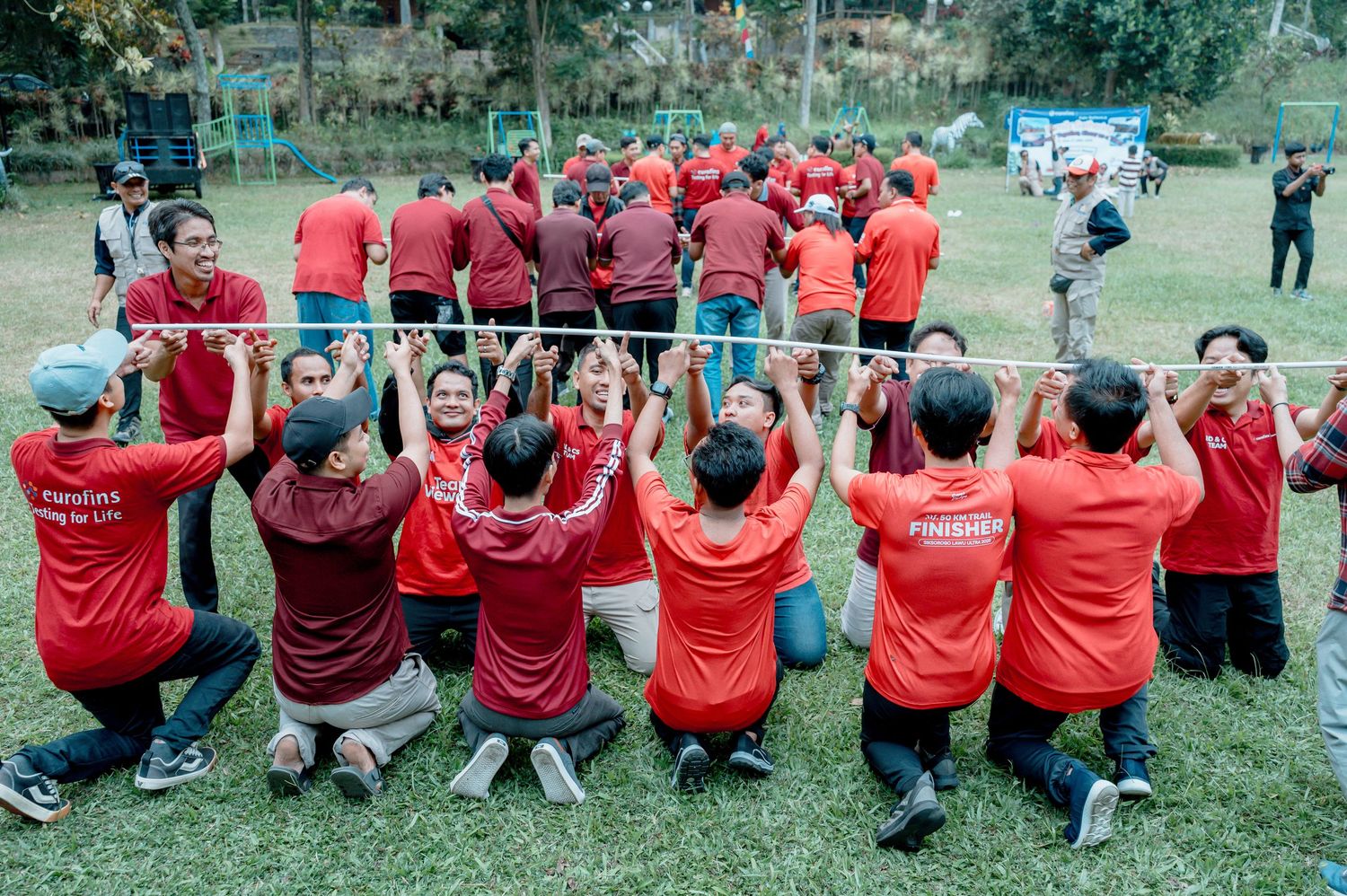 Large group in red Manulife shirts participating in outdoor team building activity, holding a metal bar together on grass.