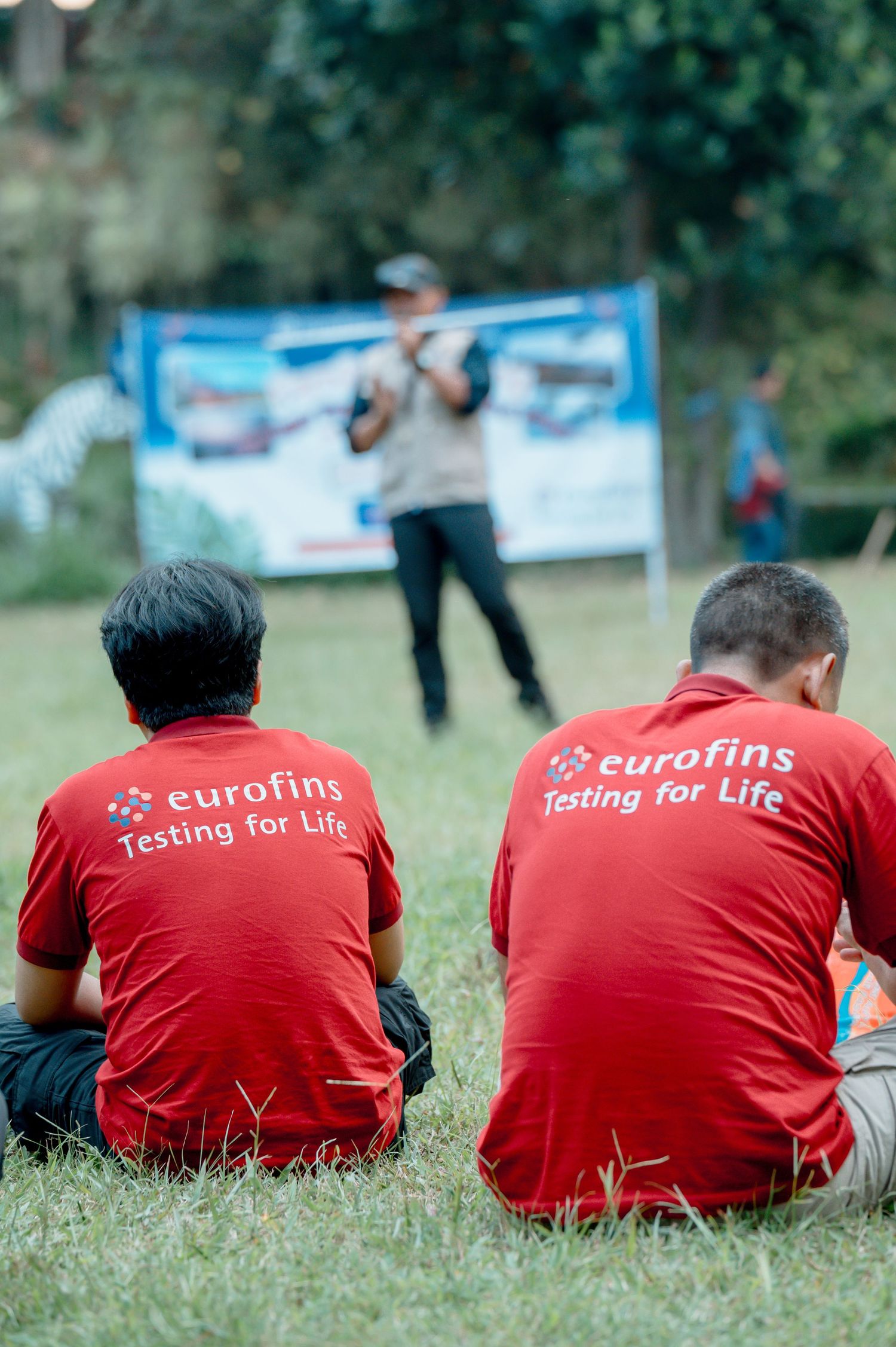 Two men in red Eurofins Testing for Life shirts sit on grass facing a presenter at an outdoor corporate event.