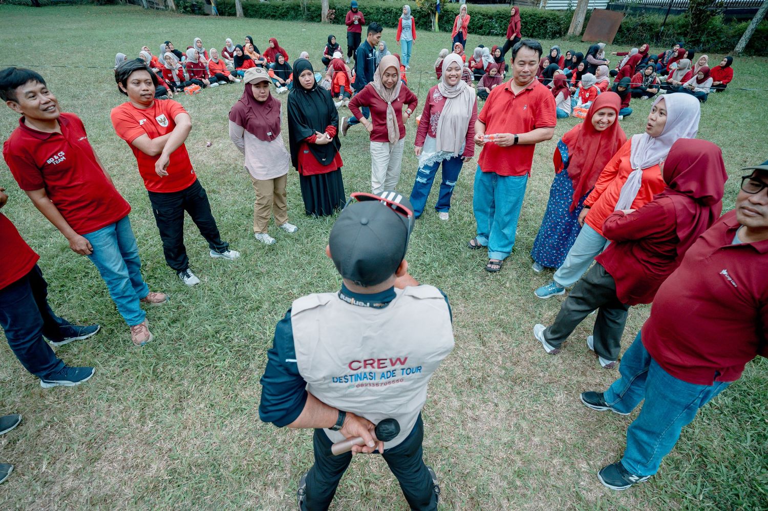 A crew member leads a team-building activity with a large group wearing red shirts outdoors on a green lawn.