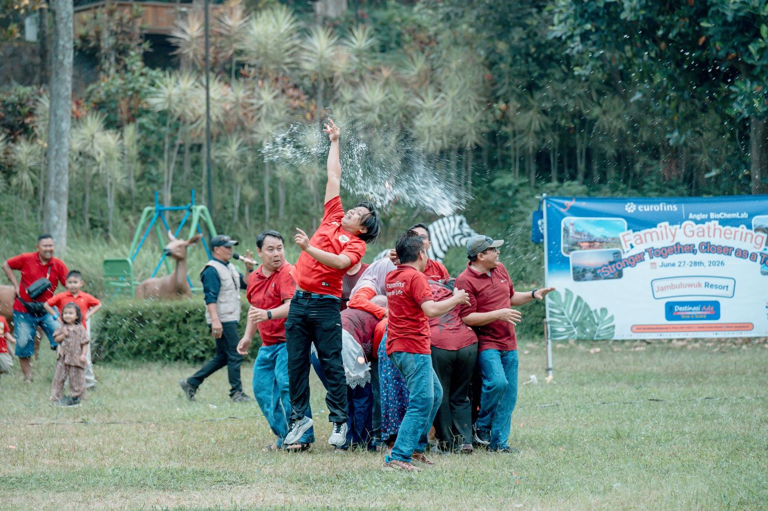 Group of people in red shirts playing outdoor team building game on green field with water splash.