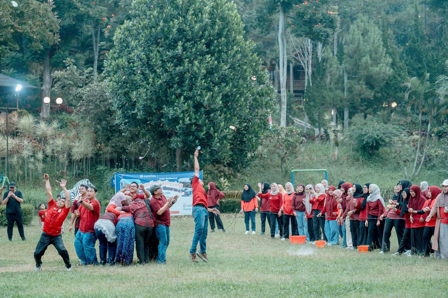 Large group of people in red shirts playing outdoor team building games on a green field surrounded by trees.
