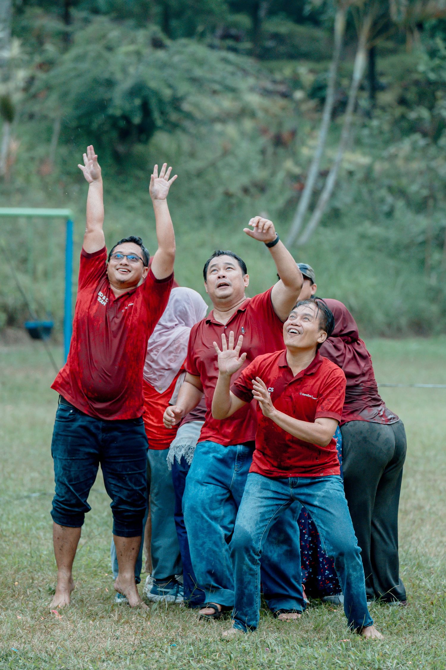 Group of men in red shirts celebrating outdoors on a grass field, raising hands in excitement during a team activity.