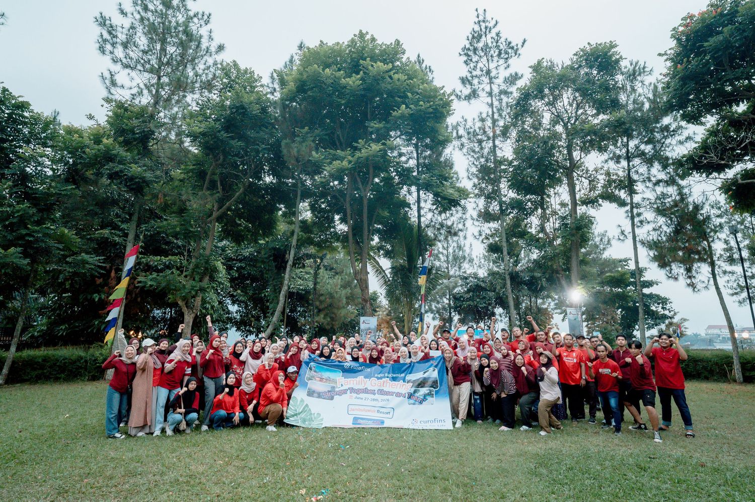 Large group of people in red shirts posing outdoors on green lawn with tall trees in background.