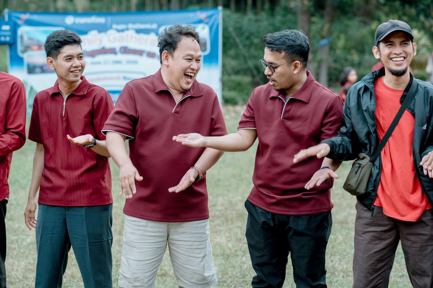 Group of men in maroon polo shirts laughing and gesturing outdoors at a team building event.