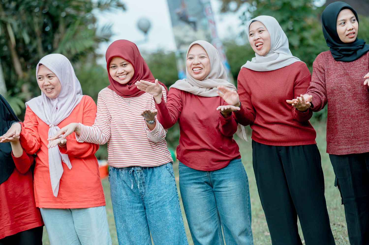 Group of women wearing colorful hijabs smiling and dancing outdoors in a park setting.
