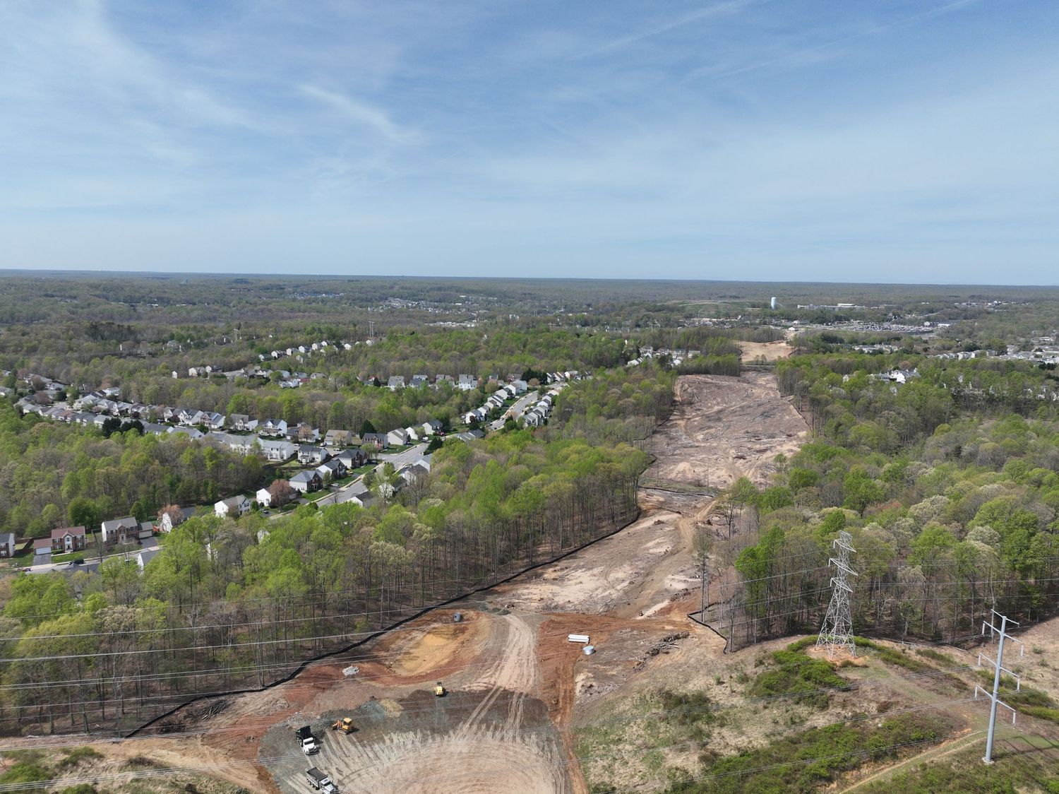 Aerial view of a large construction site with cleared red clay land near suburban neighborhoods and wooded areas.