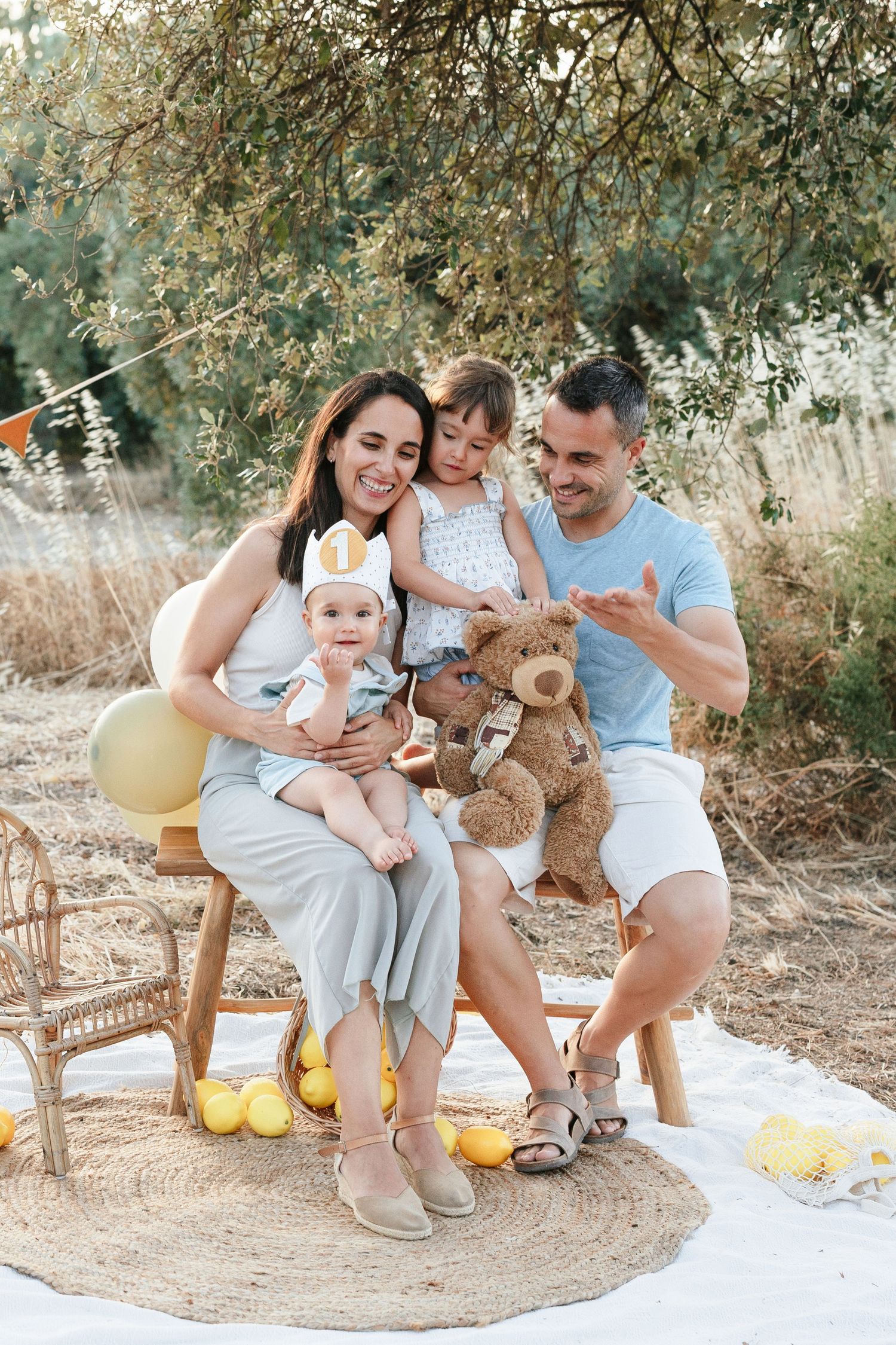 Happy family with two children sitting outdoors on a blanket with lemons, teddy bear, and balloons during a sunny day.