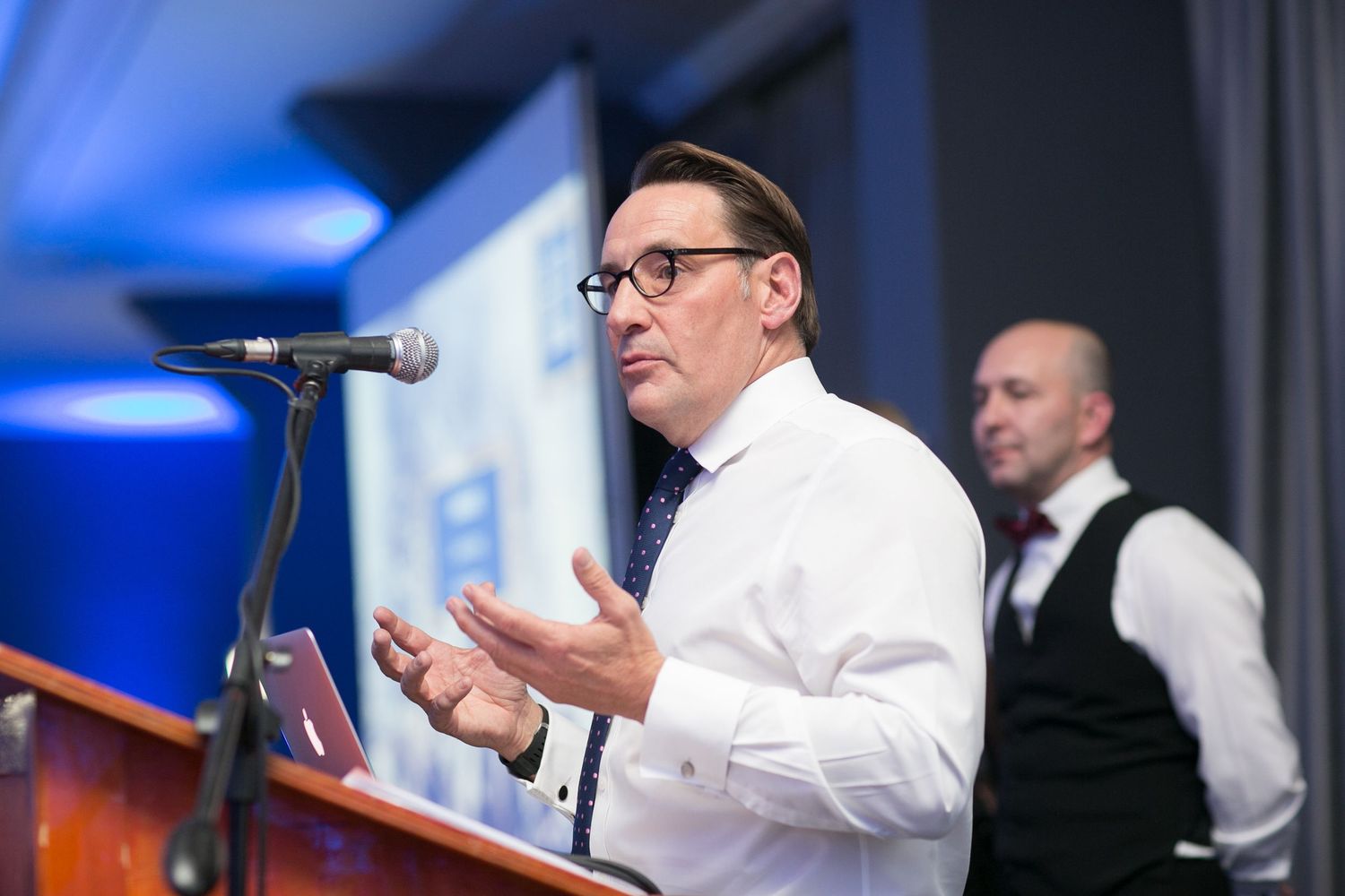 Professional speaker gesturing at podium with microphone during corporate conference event.