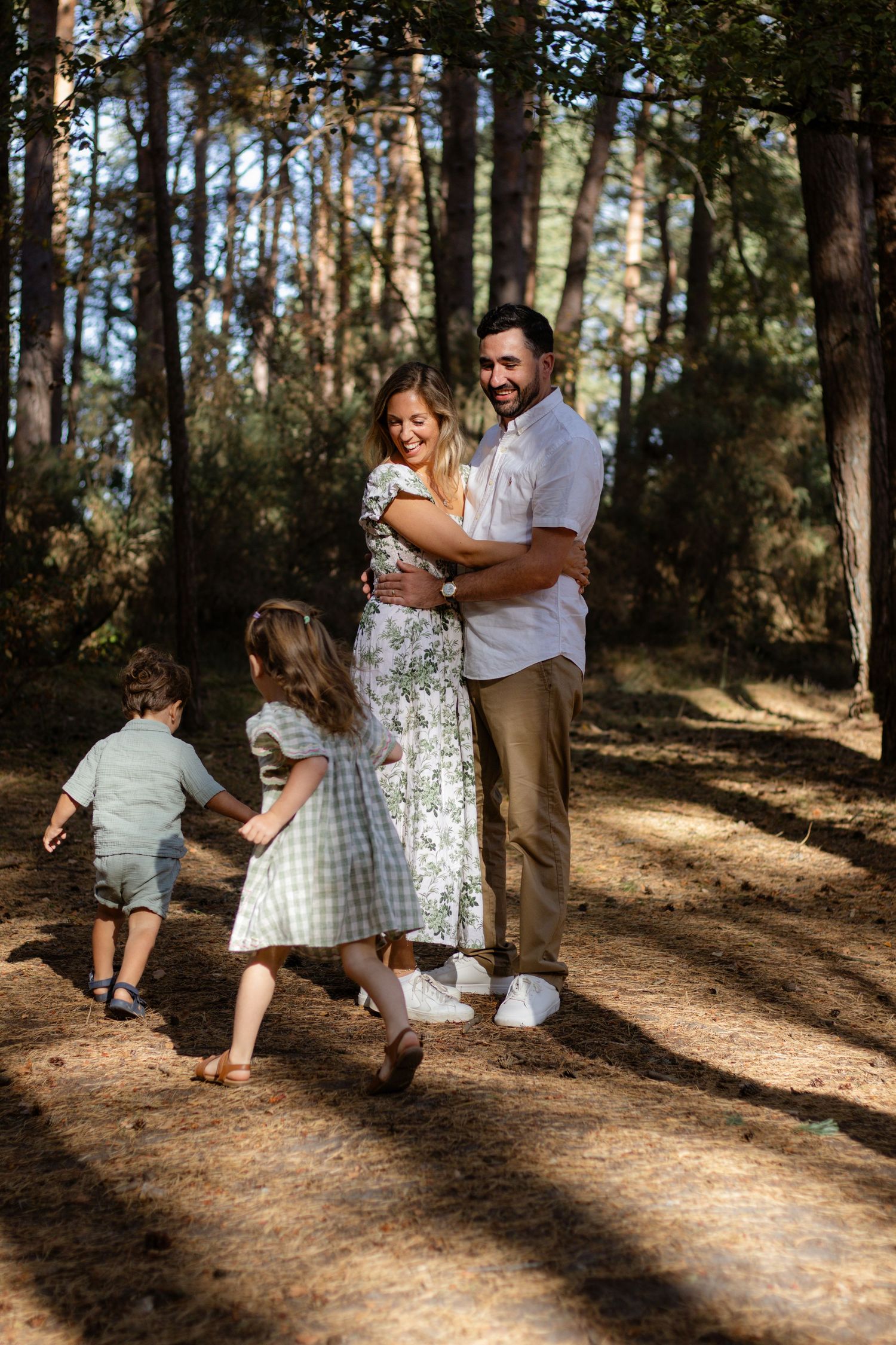 Happy family embracing in a sunlit forest, with two young children playing nearby on a pine needle path.