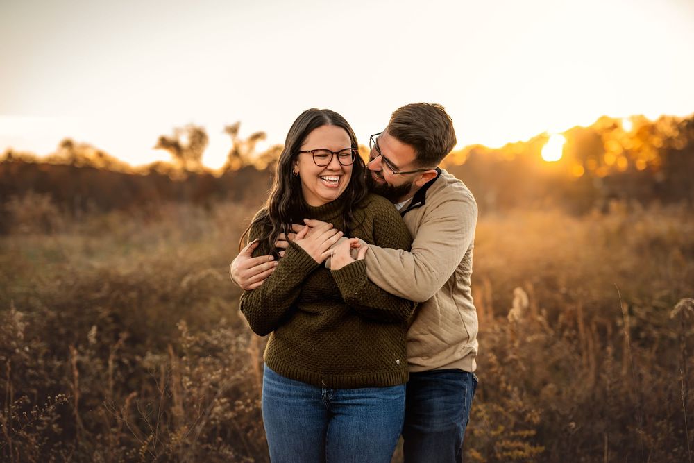 Couple embracing and laughing in golden sunset field, woman in green sweater and jeans, man in tan jacket.