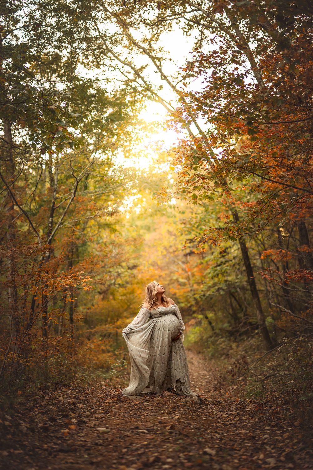 Woman in flowing gray dress walking autumn forest path with golden sunlight filtering through colorful fall foliage.