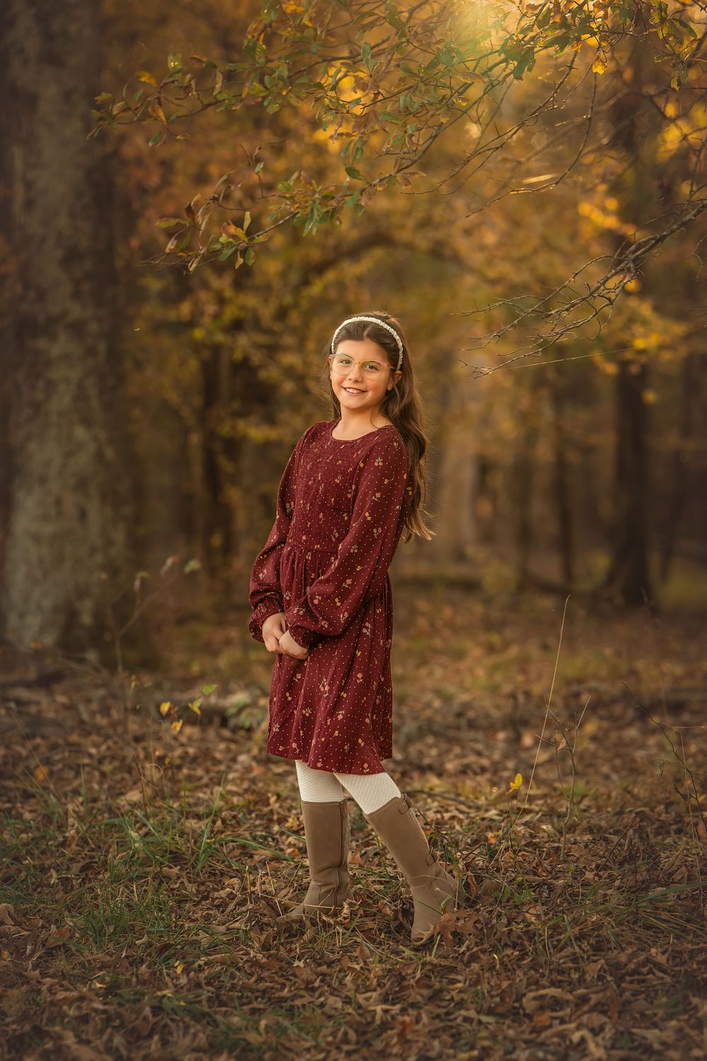 Young girl in burgundy dress and boots smiling in autumn forest with golden foliage.