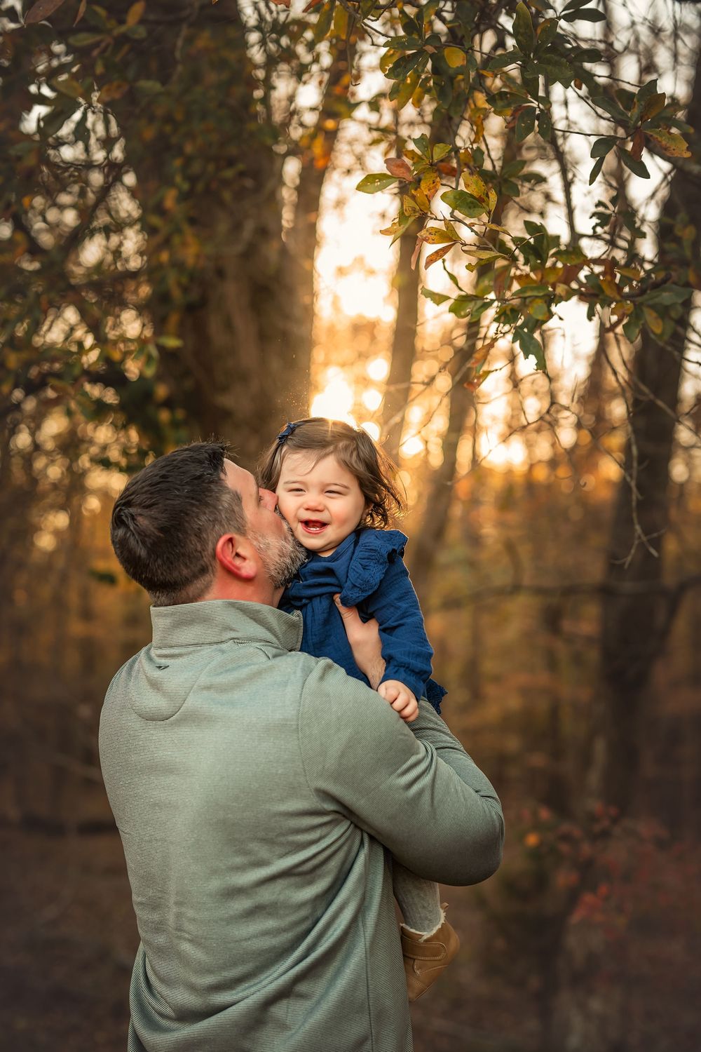 A father kisses his laughing toddler girl in a blue outfit amid golden autumn trees at sunset.