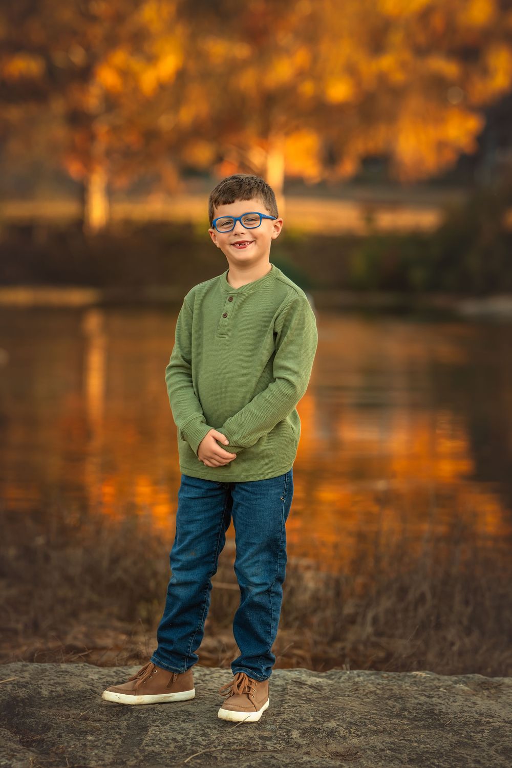 Smiling boy in green sweater and jeans with glasses poses on rocks near reflective lake with autumn trees.