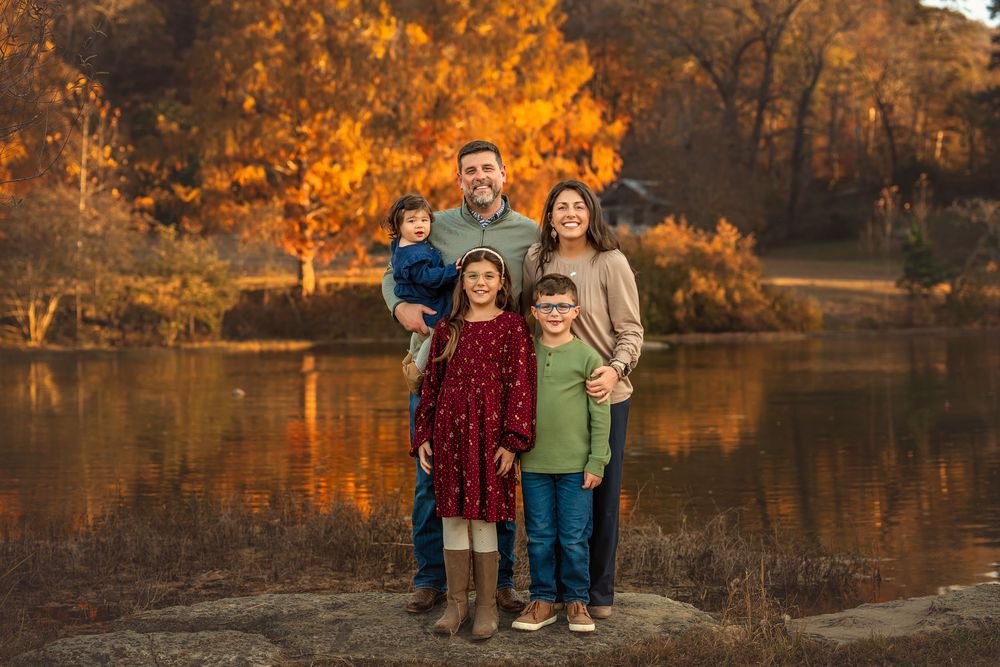 Family of five posing by a lake surrounded by vibrant autumn foliage during a fall portrait session.