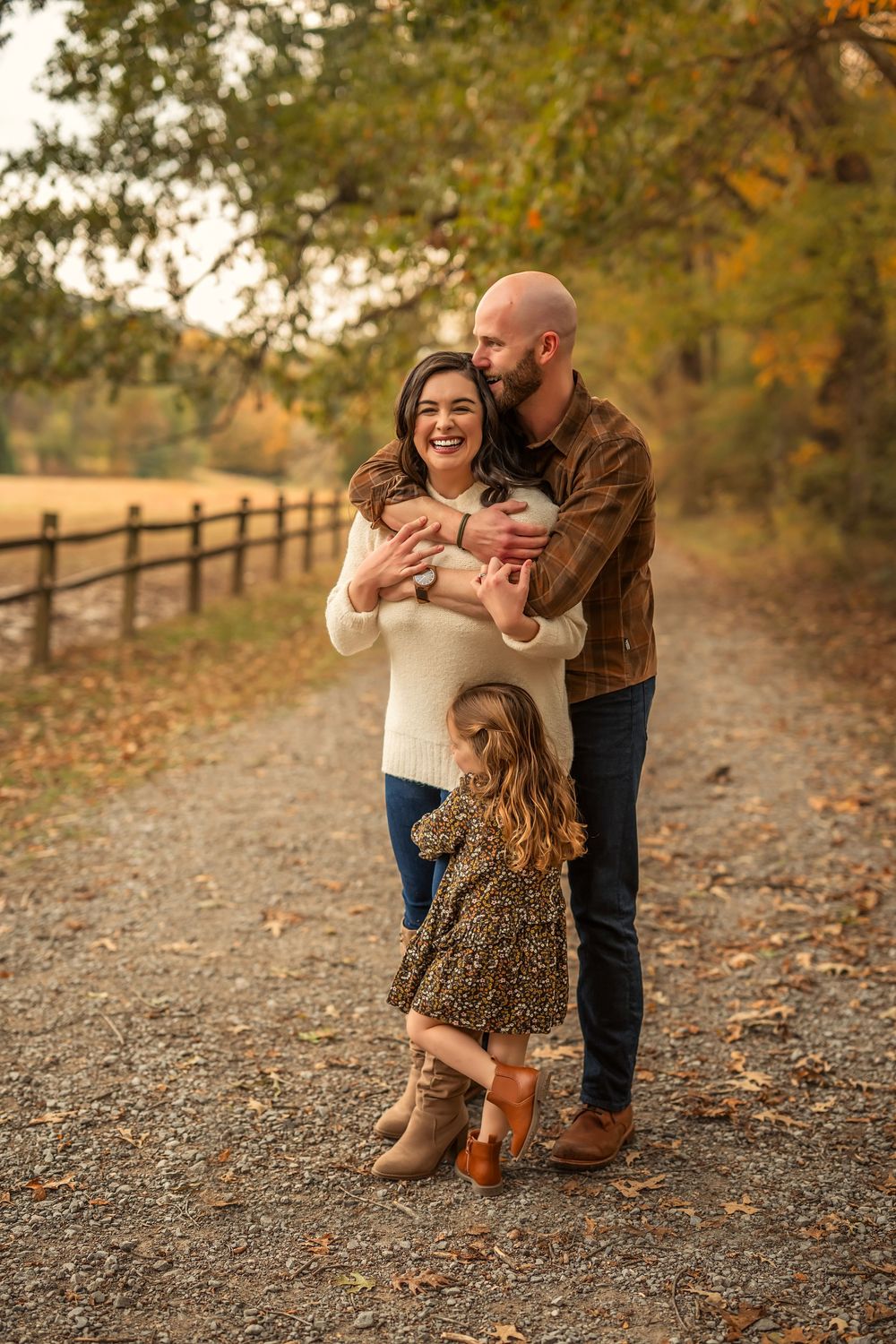 Family of three poses on a scenic autumn path, parents embracing while young daughter stands nearby.