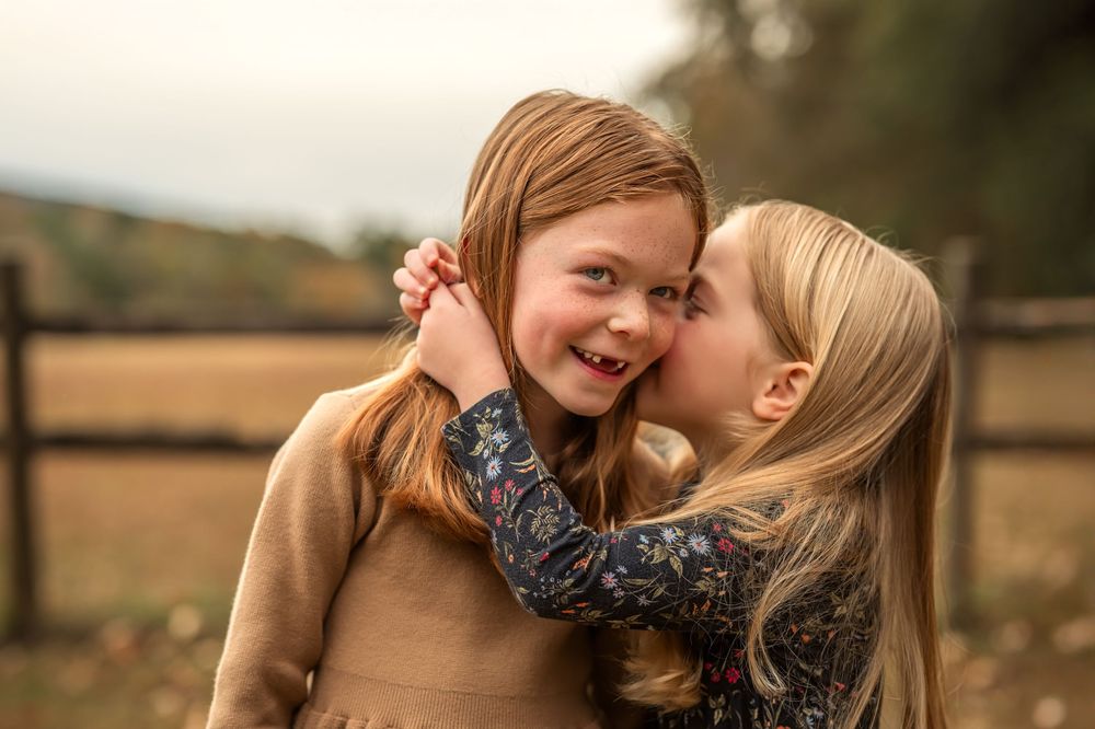 Two young girls sharing a sweet kiss on the cheek outdoors, smiling joyfully in a countryside setting.
