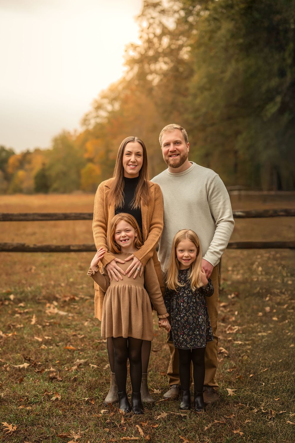 Family of four posing outdoors in autumn park, parents with two young girls holding hands, fall foliage background.
