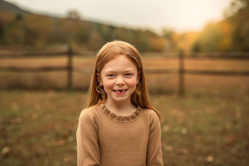 A smiling red-haired girl in a tan sweater poses outdoors on a farm with autumn foliage and a wooden fence.