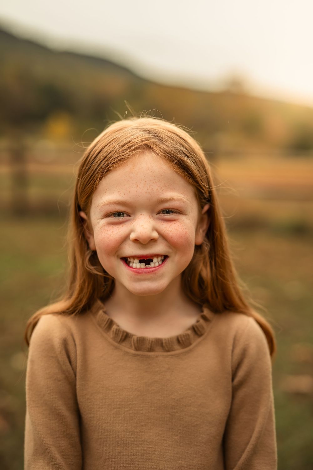 Smiling redhead girl with freckles and missing front teeth wearing brown sweater outdoors in golden hour light.