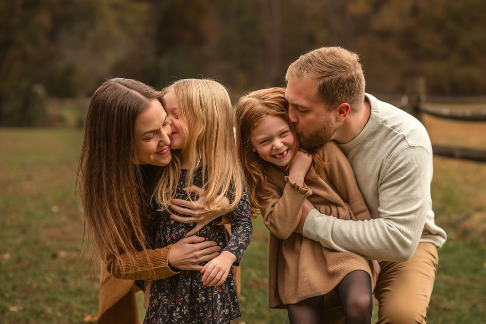 Happy family of four sharing a joyful moment outdoors in autumn, parents kissing and hugging two young daughters.