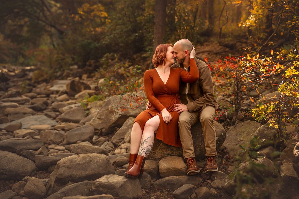 Couple sharing a romantic moment on rocky riverbed surrounded by vibrant autumn foliage in a forest setting.