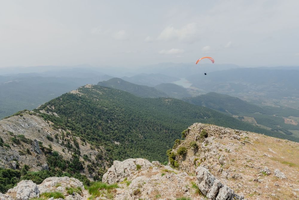 Paraglider soaring over lush green mountain ridges under a hazy sky in a scenic aerial adventure.