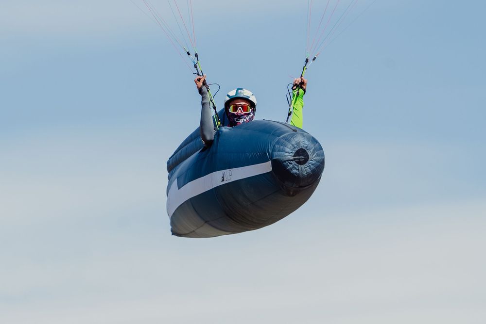 Paraglider soaring through a clear blue sky with a dark oval-shaped wing and colorful harness equipment.