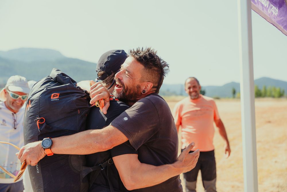 Two men sharing a joyful hug outdoors at a sunny event with mountains in the background.