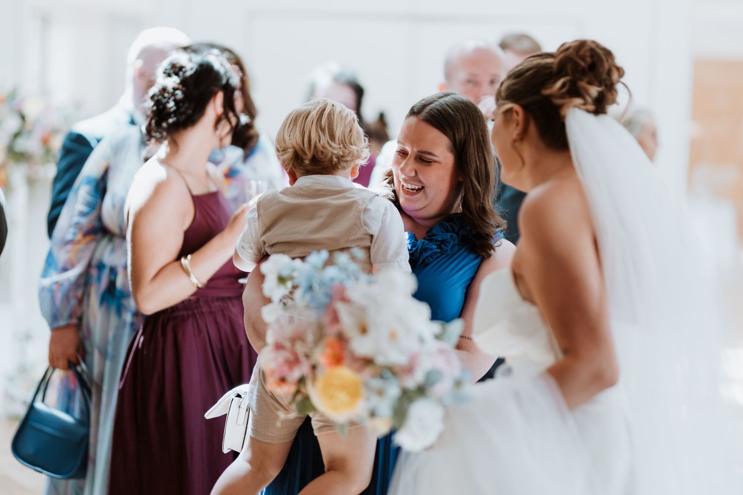 Bride in white gown receives guests at wedding reception, holding colorful bouquet while woman in blue dress holds young child.