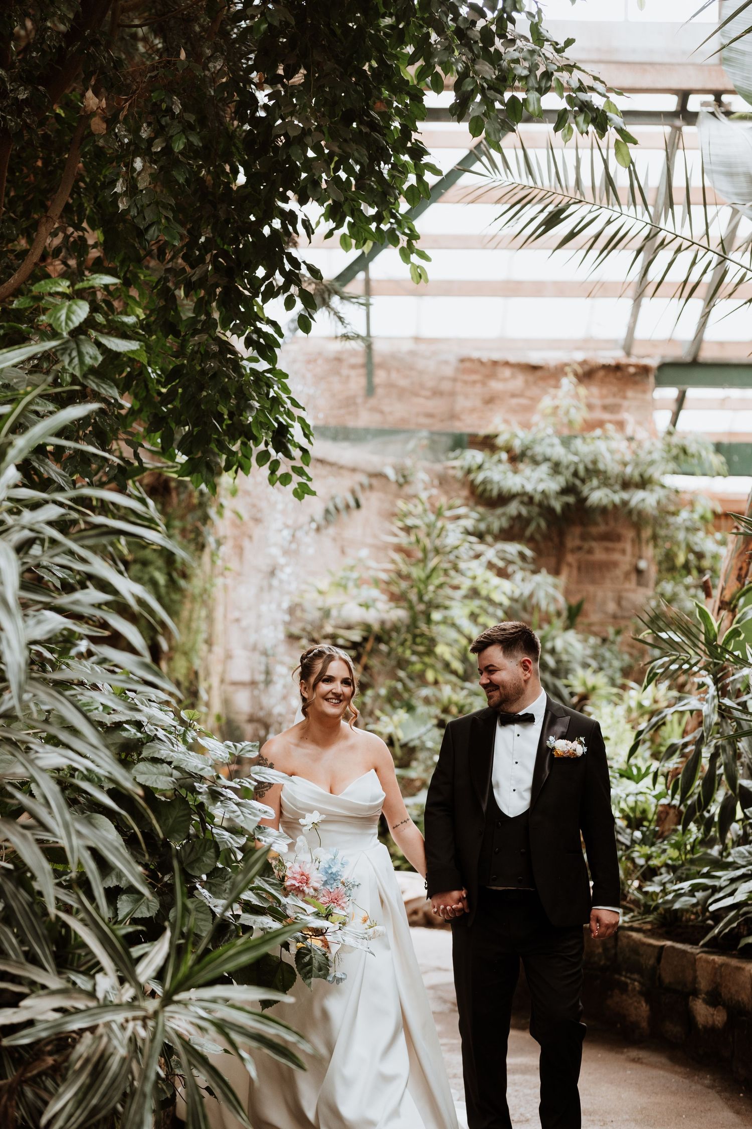 Bride and groom walk hand-in-hand through a lush greenhouse garden, she in a white gown, he in a black tuxedo.