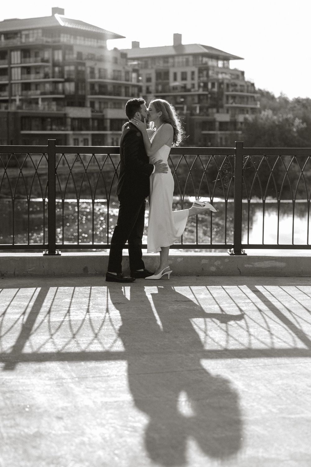 Black and white photo of newlyweds kissing on a sunny bridge with their shadows on the ground.