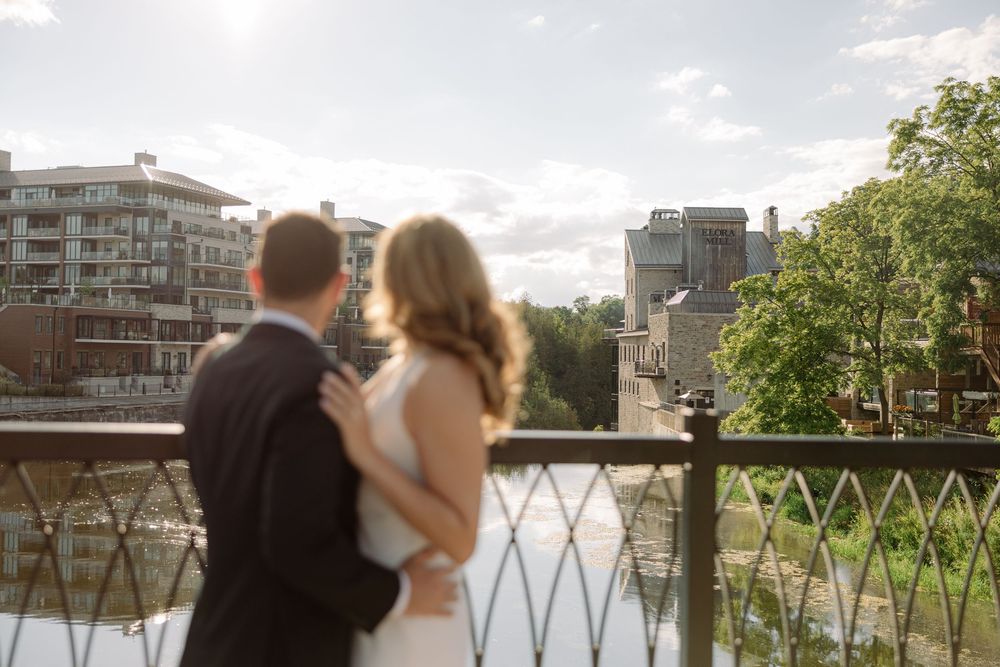 Bride and groom embracing on a decorative bridge overlooking a river and urban skyline.