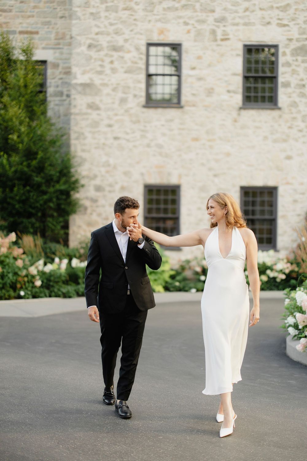 Groom in black suit kissing bride's hand while she smiles in white halter dress outside stone building.