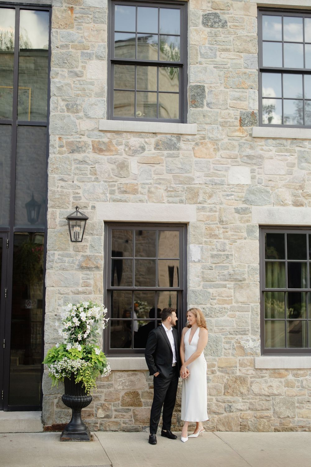 Elegant couple holding hands and smiling at each other in front of a stone building with floral arrangement.