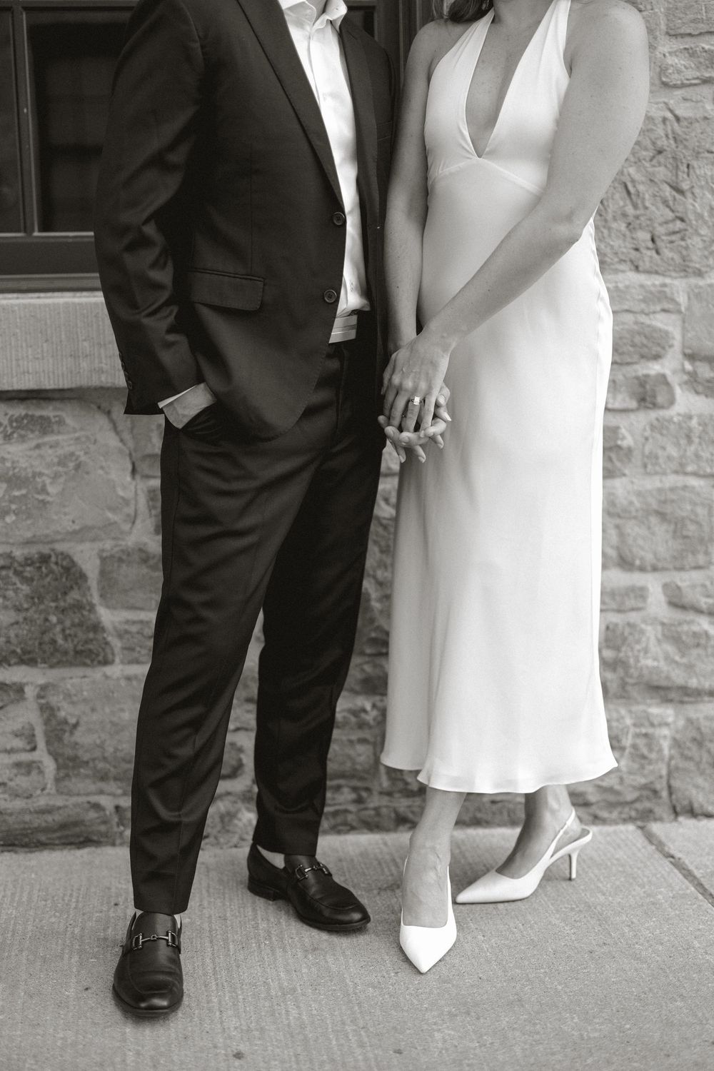 Black and white photo of bride and groom holding hands in front of a stone wall, showing wedding rings.